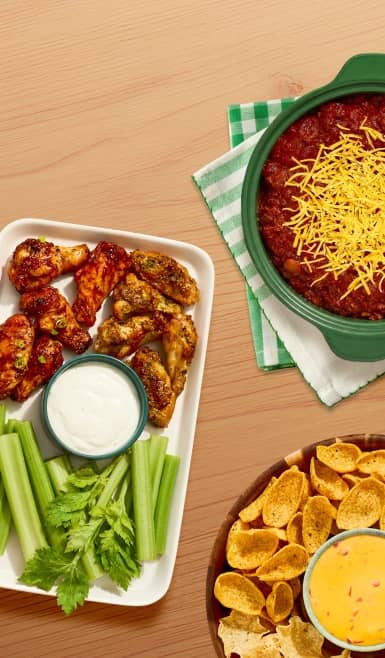 Two different kinds of chicken wings, celery sticks, and creamy dressing next to chili topped with cheese and corn chips with queso dip.