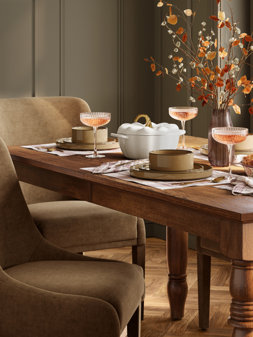 Wood dining table set with champagne coupes, plates and a white ceramic serving dish, with upholstered chairs and a vase of autumn branches.