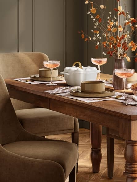 Wood dining table set with champagne coupes, plates and a white ceramic serving dish, with upholstered chairs and a vase of autumn branches.