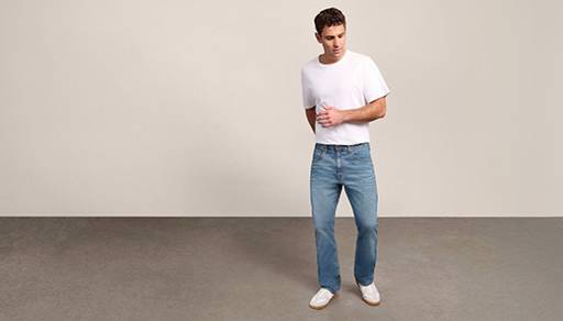 A man wearing bootcut jeans and white tee shirt is pictured on a beige backdrop.