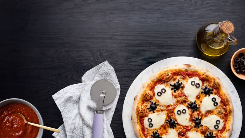 A pizza with mozzarella cheese ghosts with sliced black olive eyes next to pizza sauce, a pizza wheel, olive oil and sliced olives.