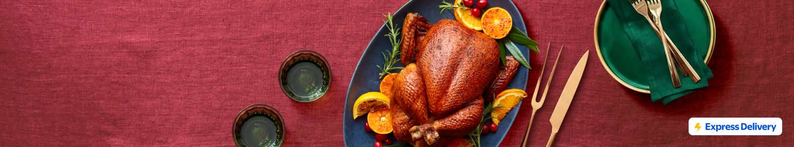 Express Delivery. A roasted turkey on a platter with orange slices and wedges, herbs, and cranberries against a burgundy background.
