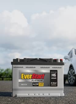 A car battery on pavement, with a car in the background.