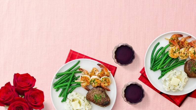 Two plates with steak, shrimp, mashed potatoes and green beans next to two glasses of red wine and red roses.