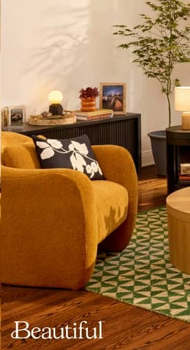 A mustard yellow upholstered chair sits at the edge of a green and cream rug. The living room is set with a wood console and end table with decor.