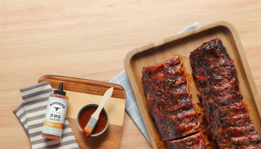 A cutting board with a bottle and bowl of Kinder’s BBQ sauce honey hot next to a tray with two racks of cooked, sauced ribs.