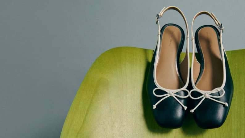 A pair of black slingback flats with white trim and bow details rests side by side on a green surface.