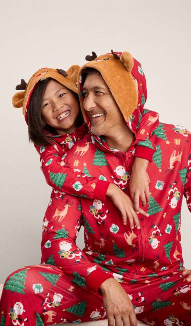 A father and child wearing matching reindeer onesie pajamas are pictured on a beige backdrop.