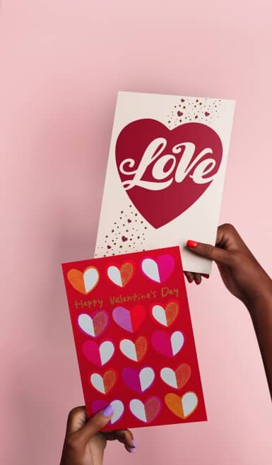 Two hands hold two Valentine’s Day cards. One has a red heart and says “Love” and the other is a red card with multi-color hearts.