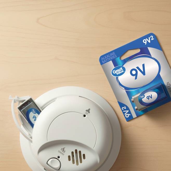 A smoke detector with the battery door open and a battery sticking out. A pack of Great Value nine volt batteries.