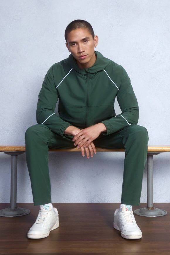 Man sitting on a bench wearing a forest green zip-up active hoodie, green sweatpants and white sneakers in a studio.