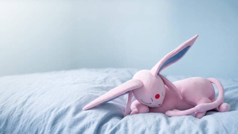 Pokemon Espeon plushie in a sleeping pose on a bed.