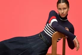 Woman posing in a chair wearing a navy turtleneck with red and white stripes, pleated denim skirt and black belt.