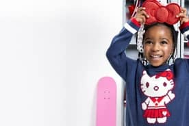 Smiling girl in navy Hello Kitty sweater holds a red bow purse above her head in front of lockers and a pink skateboard.
