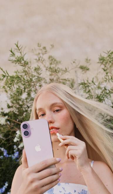 A person holding an iPhone with a pink case and applying lip gloss.