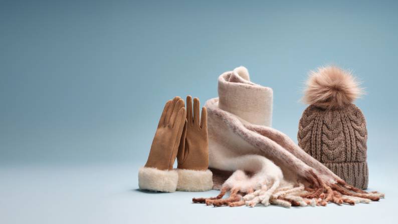 Women’s tan faux suede gloves, tan striped blanket scarf and a brown cable knit pom beanie are shown.