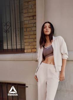 A young woman is shown leaning against a building wearing a Reebok sports bra, zip-up jacket and matching sweats.
