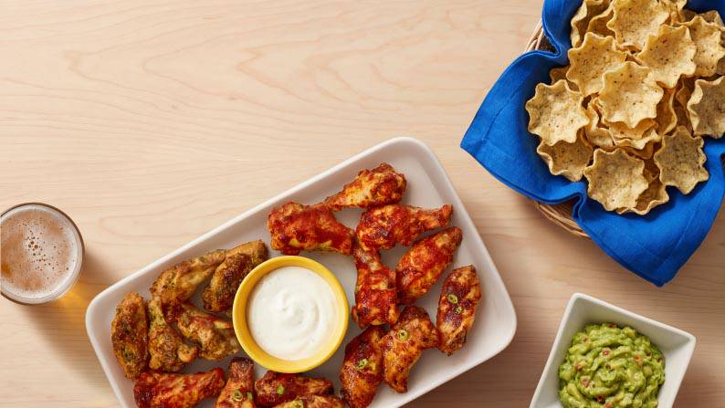 A beverage, a plate of wings with dip, a bowl of guacamole, and a bowl of tortilla chips with a blue napkin.