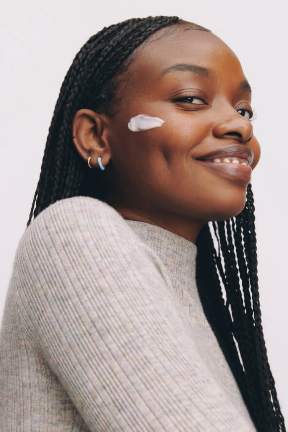 Smiling adult with long braids in a ribbed sweater shows a swipe of face cream on one cheek.