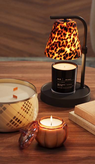 Candles on a wooden table with a lit pumpkin candle, a large patterned candle and a black candle warmer with leopard print shade.