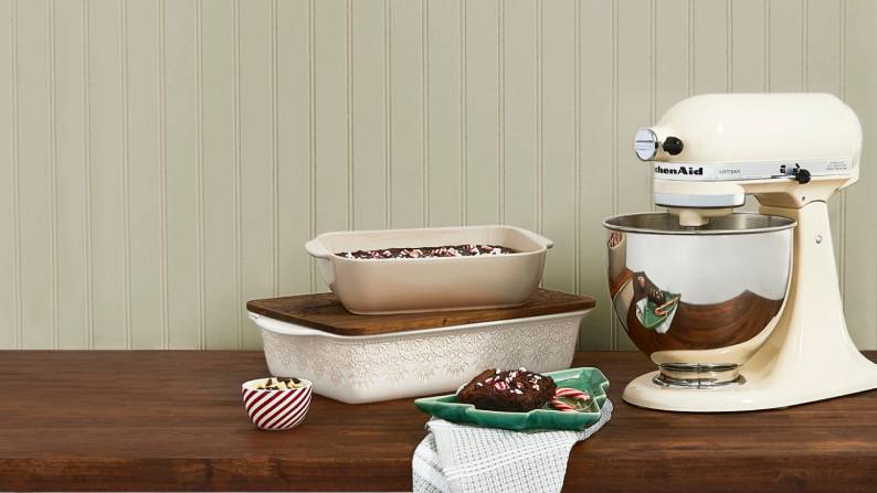 A cream-colored stand mixer, bakeware and treats sit on a wood countertop against sage wainscot walls.