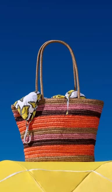 A striped woven tote bag in shades of orange, pink, brown and red with long handles and a scarf tucked inside is set against a blue sky background.