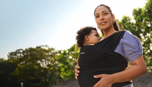 A mother in a black baby wrap carrying an infant, next to trees.