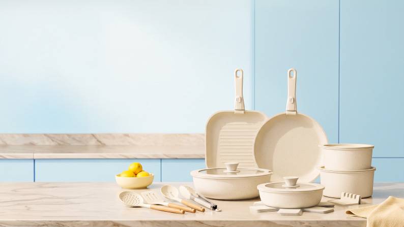 Cream cookware set with pans, lidded pots, utensils and a bowl of lemons on a marble countertop.