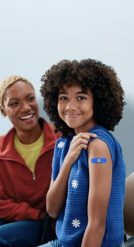 A little girl with a band aid on her arm, smiling and sitting alongside another woman.