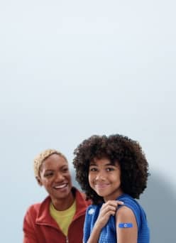 A little girl with a band aid on her arm, smiling and sitting alongside another woman.