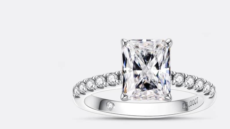 A square cut diamond ring with an embellished silver band is pictured on a light grey backdrop.