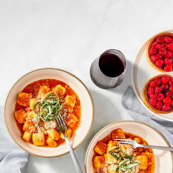 Two bowls of gnocchi in red sauce topped with fresh herbs and grated cheese next to a glass of wine and two raspberry tarts.