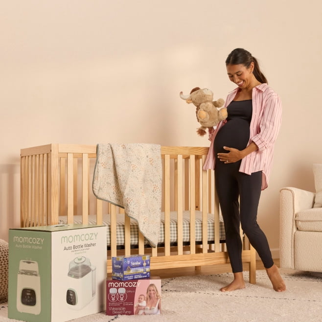 Momcozy baby products beside a crib as a pregnant adult stands holding a stuffed bear.