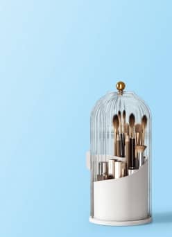 Makeup brushes and cosmetic containers in a white organizer covered by a clear cloche with a gold knob.