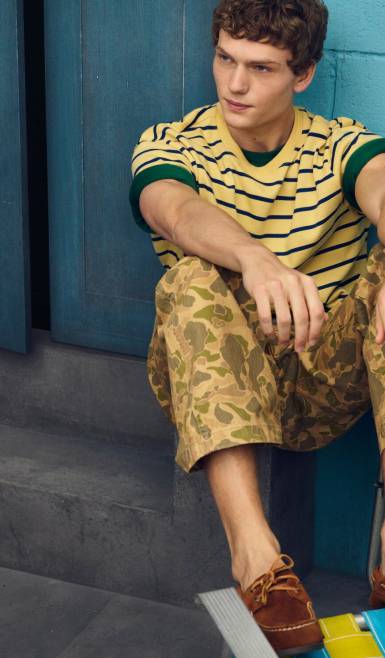 Man in a yellow-and-navy striped tee, camo-print pants, and brown loafers, sitting on steps against a blue wall.