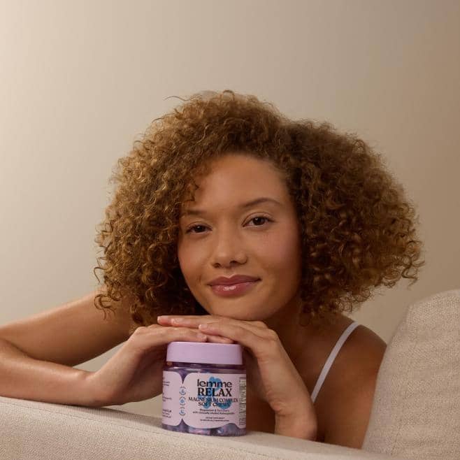 Adult with curly hair leans on a surface, holding a lavender lemme RELAX hair mask jar in front.