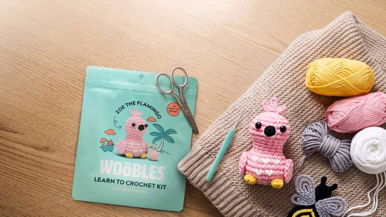 Wobbles Zoe the Flamingo learn to crochet kit with finished flamingo toy, yarn balls and tools on a wooden table.