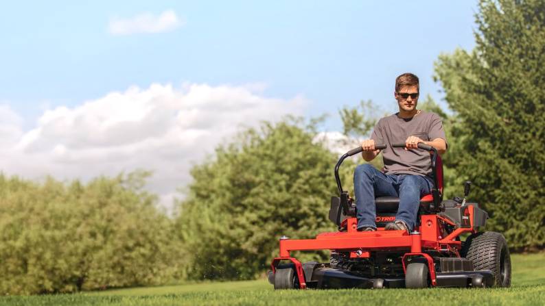 A person driving a ride-on mower in front of trees.