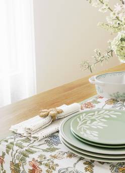 A wood table decorated with a floral printed runner, green table settings, gold flatware, cloth napkins, and a vase of flowers.