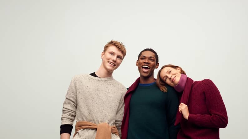 Three adults in cozy sweaters stand close together smiling, with one resting their head on another shoulder.