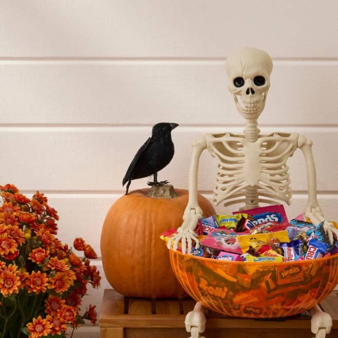 Skeleton decor holds a large orange bowl filled with assorted wrapped Halloween candy beside a pumpkin and orange flowers.
