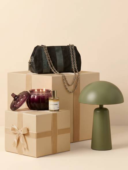Black textured handbag with chain straps on gift boxes with lit dark glass candle, fragrance bottle and green modern table lamp.