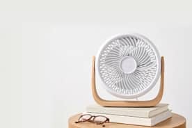 A white fan on top of a stack of books and a wood table.