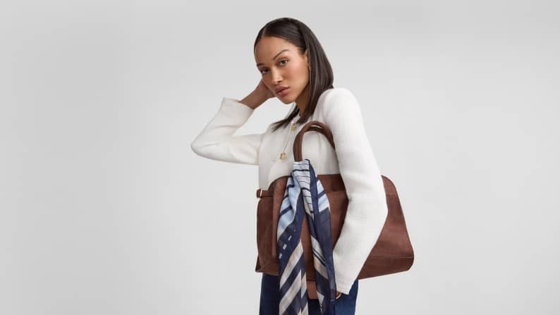Woman wearing a white jacket, jeans and holding on to a brown satchel handbag with a navy striped scarf tied around handle.