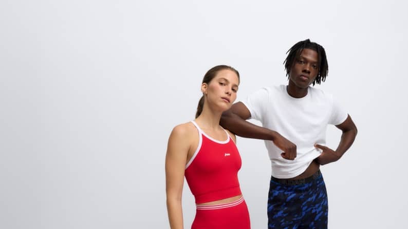 Two adults pose in athletic wear, one in a red sports set and one in a white shirt with blue patterned shorts.