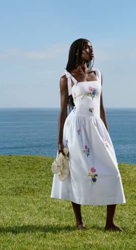 Young adult woman in white midi dress with floral embroidery holding white bow sandals by the sea.
