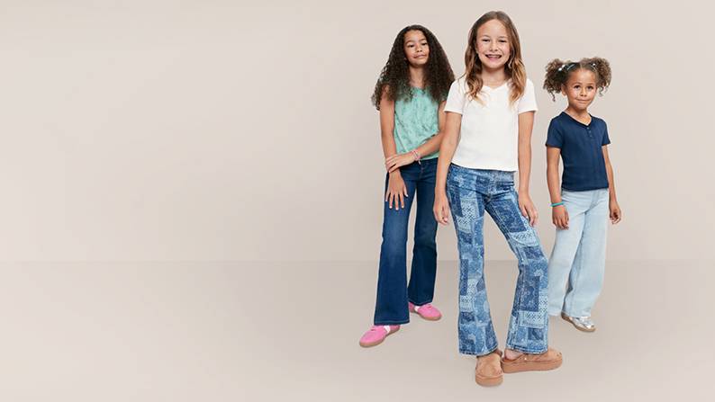 Three girls wearing jeans and tee shirts are pictured on a beige backdrop.
