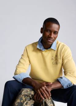 Man seated wearing a pale-yellow sweater layered over a blue button-up shirt with navy pants, holding a camo-print piece of clothing.