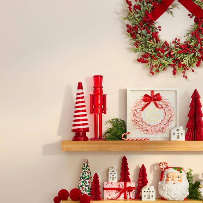 Christmas wreath above shelves with red and white trees, nutcracker, wrapped gifts, Santa figurine and small decorative houses.
