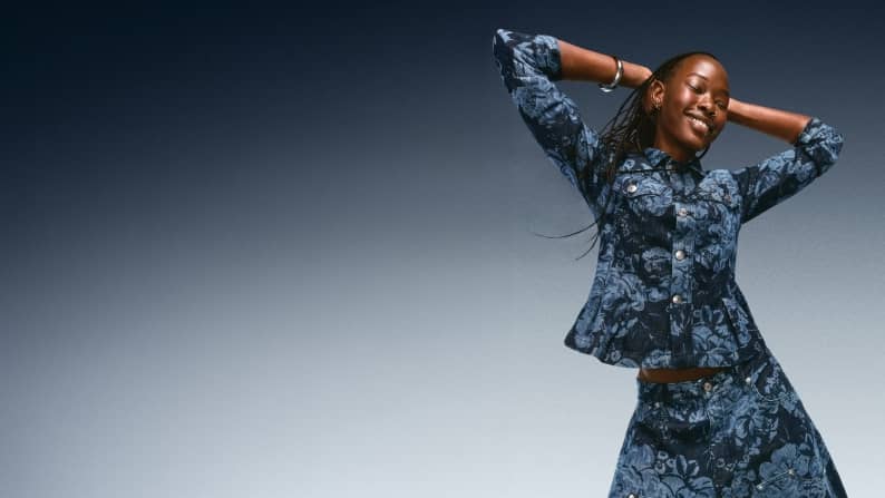 Woman posing in a matching dark blue floral-patterned denim peplum jacket and matching jeans with hands behind her head.